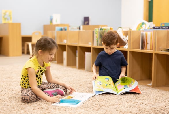 Two children sitting on the floor reading books in a library.