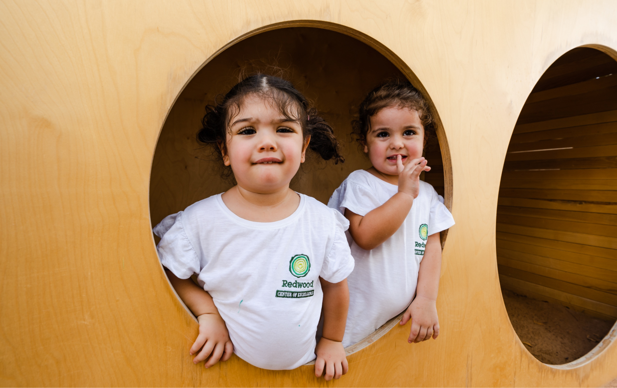 Two young children smiling in circular cutouts of a wooden play structure, wearing white t-shirts with logos.