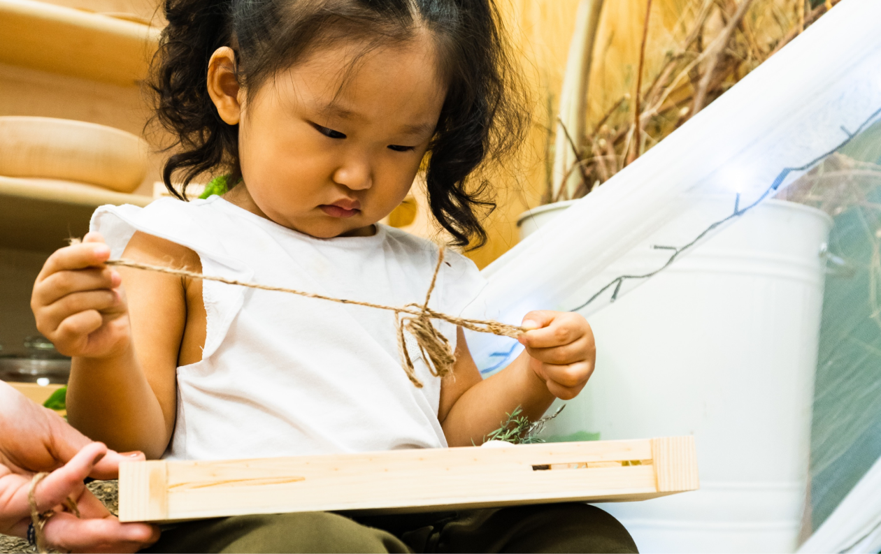 A young girl concentrating on tying a string around a small wooden frame, surrounded by plants.