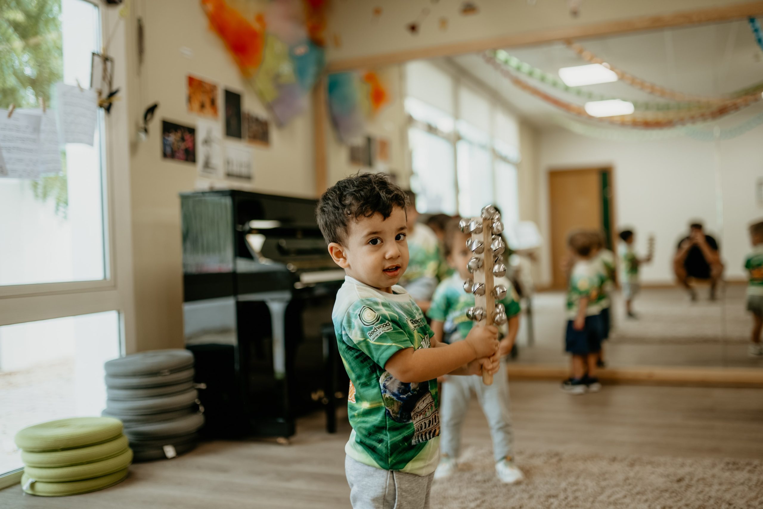 Toddler boy in a green shirt playing with a musical triangle in a classroom with other children in the background.