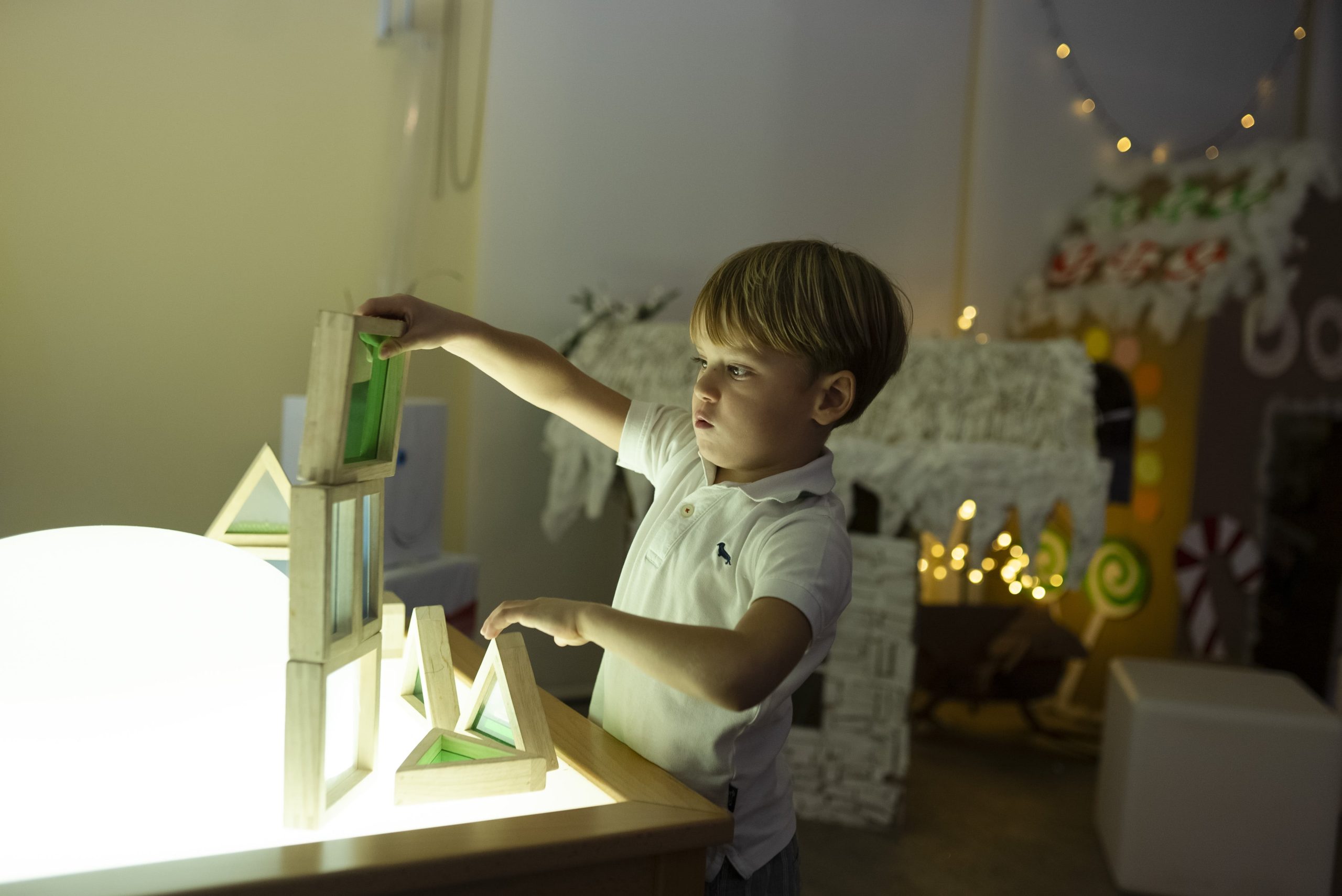A young boy plays with a wooden block set on a light table in a dimly lit room decorated with fairy lights.
