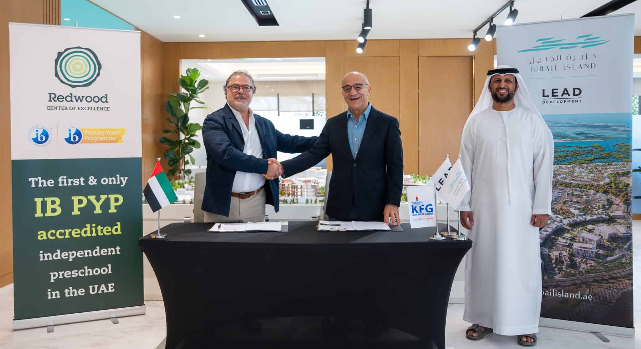 Three men stand in a room with two shaking hands across a table. Banners for Redwood and Jubail Island are displayed behind them.